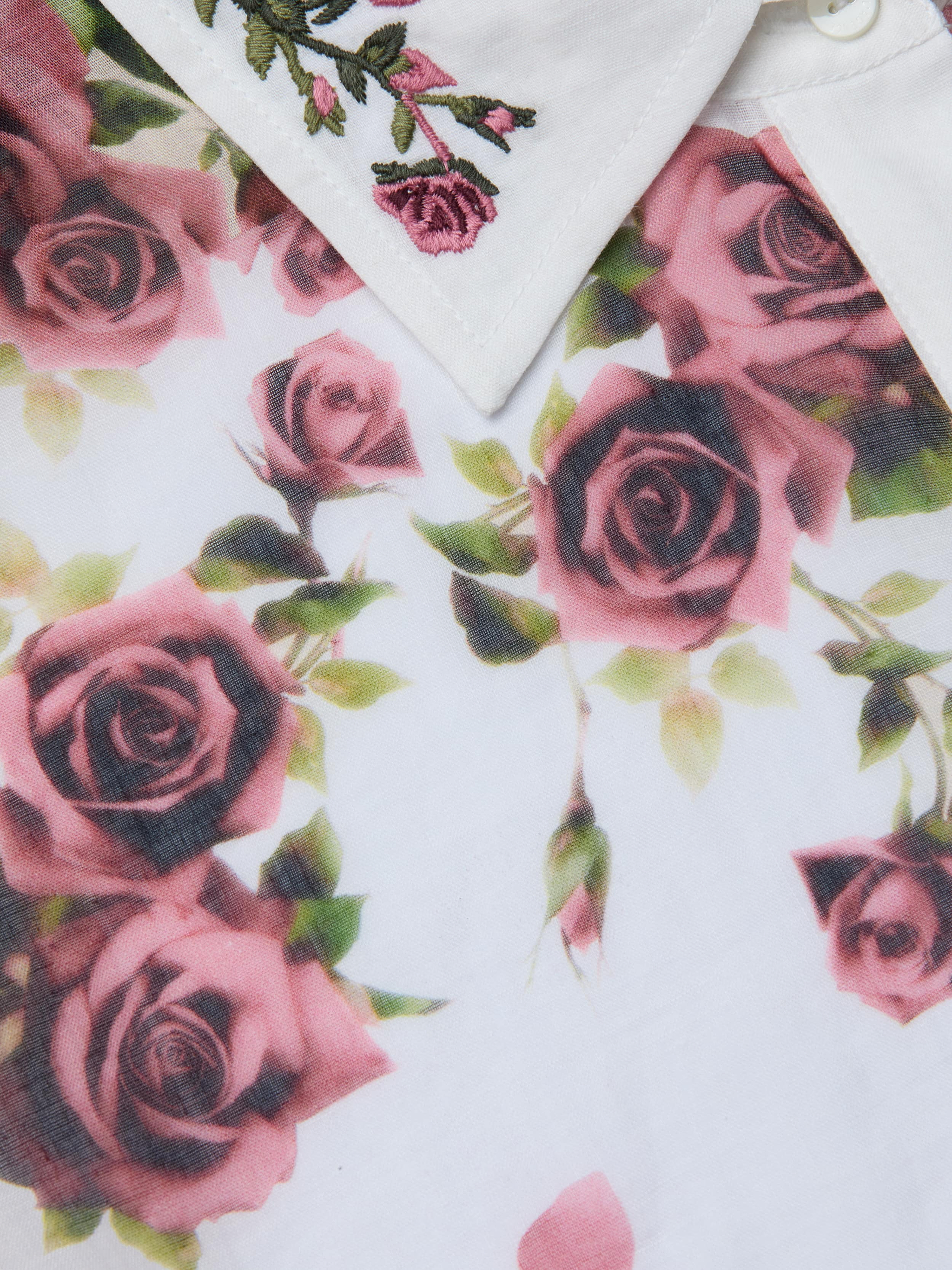 Close-up of the Philomena Blouse—a white button-down shirt with a collar, showcasing embroidered pink roses and green leaves, complemented by a rose print pattern with pink roses and green stems on semi-sheer Ramie fabric.
