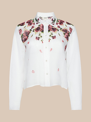 The Philomena Blouse is a white Ramie button-down shirt with a collar, adorned with pink rose and green leaf prints across the shoulders and upper chest, plus scattered pink petals on the front.