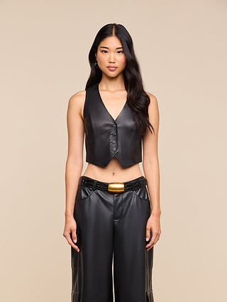 A woman with long black hair wears the Jianna Leather Vest, paired with matching high-waisted pants, standing against a beige background—a striking look inspired by Fall 2026 fashion.