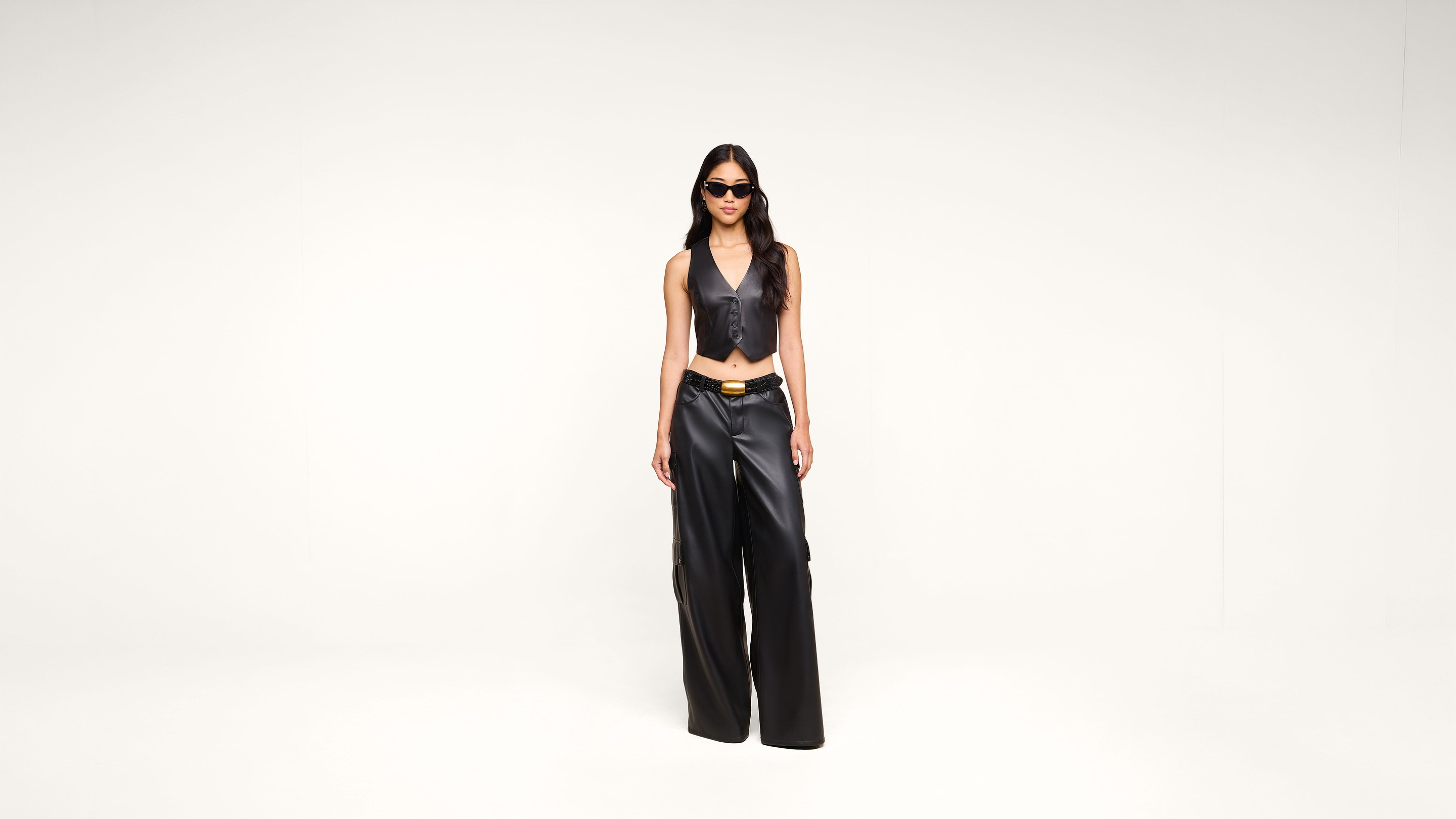 A woman wearing sunglasses, a black leather vest, and matching wide-leg pants stands confidently against a plain white background.