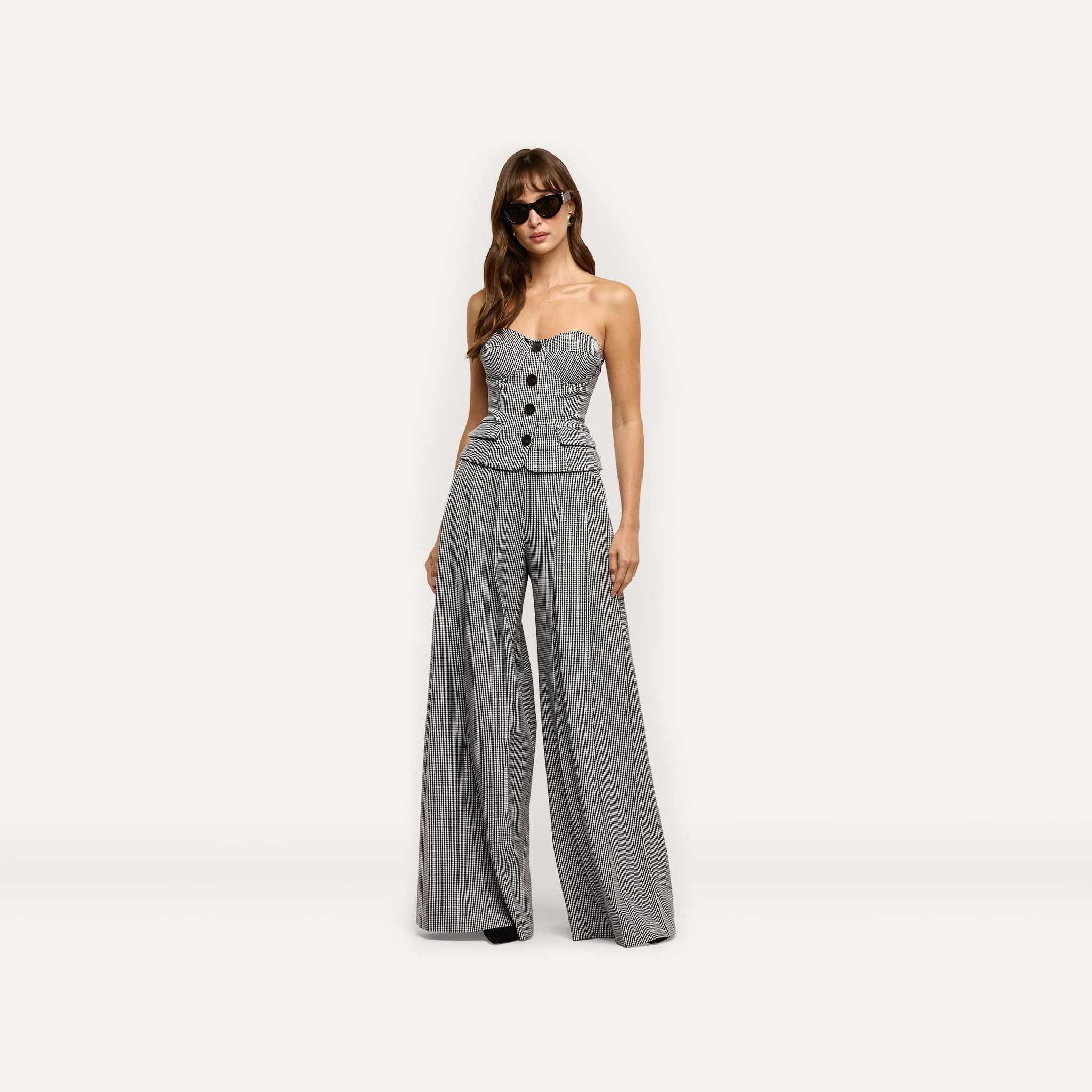 A person wears a black and white houndstooth Genevieve Corset Top featuring a strapless, button-front design with a peplum hem, paired with matching high-waisted wide-leg trousers.