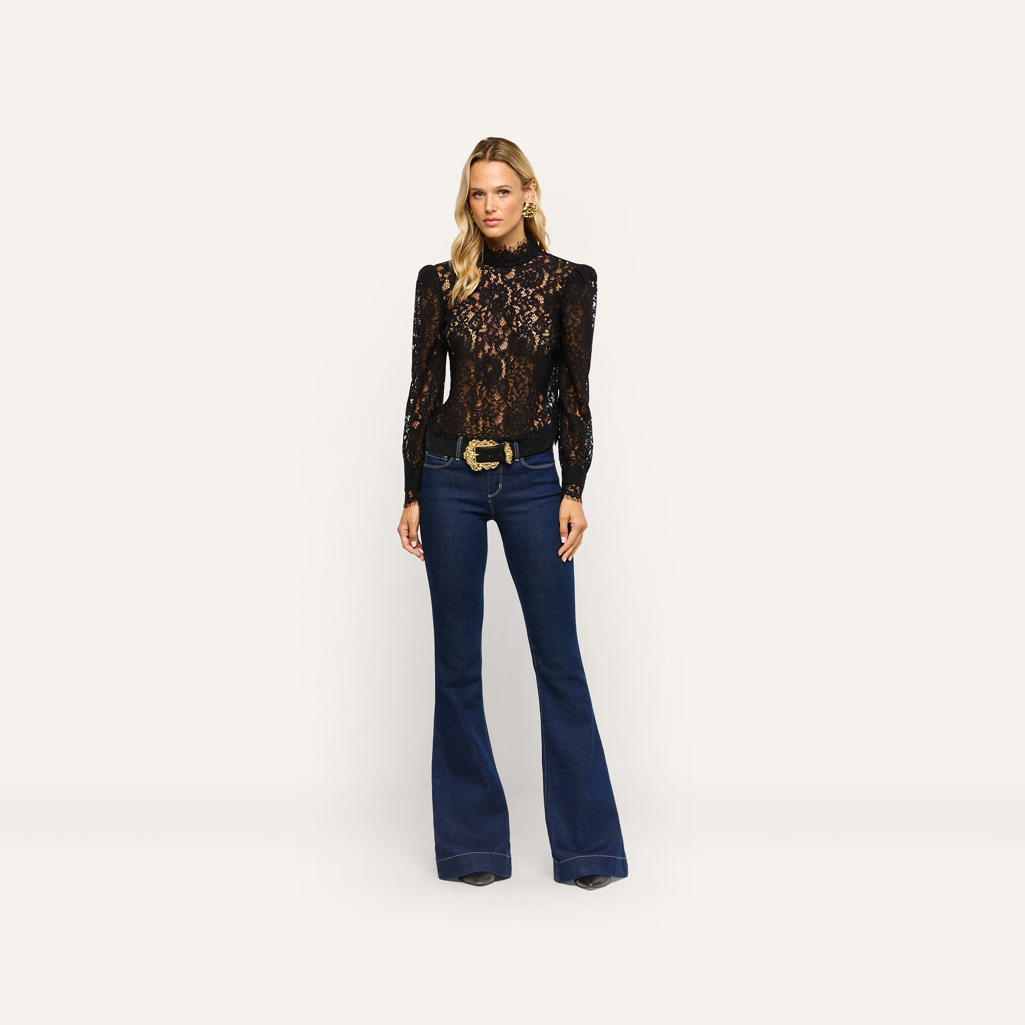 A woman wearing an Amarenthe Lace Blouse in black paired with dark wash flared jeans featuring a large gold belt buckle.