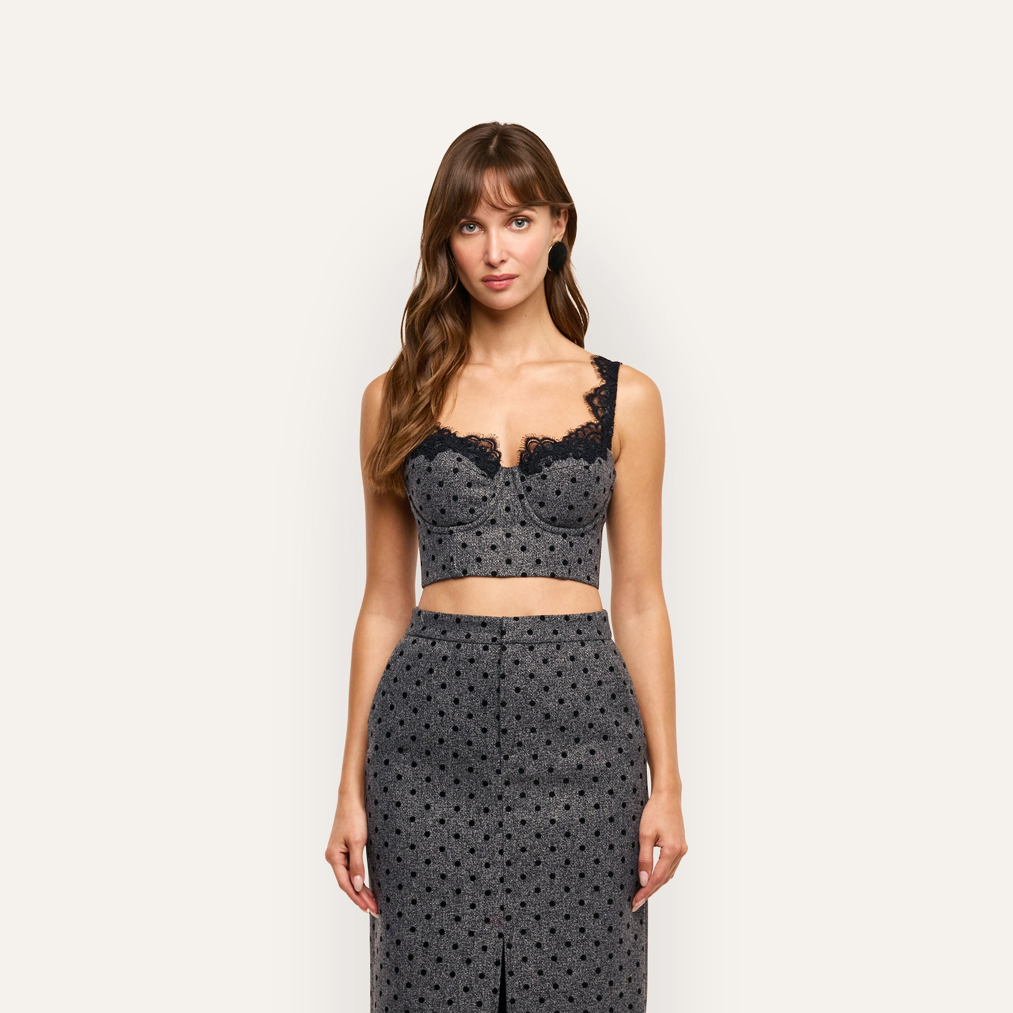 A model wears an Oriane Bustier Top in grey with polka dots, built-in underwire, and black lace trim, paired with a matching midi-length skirt.