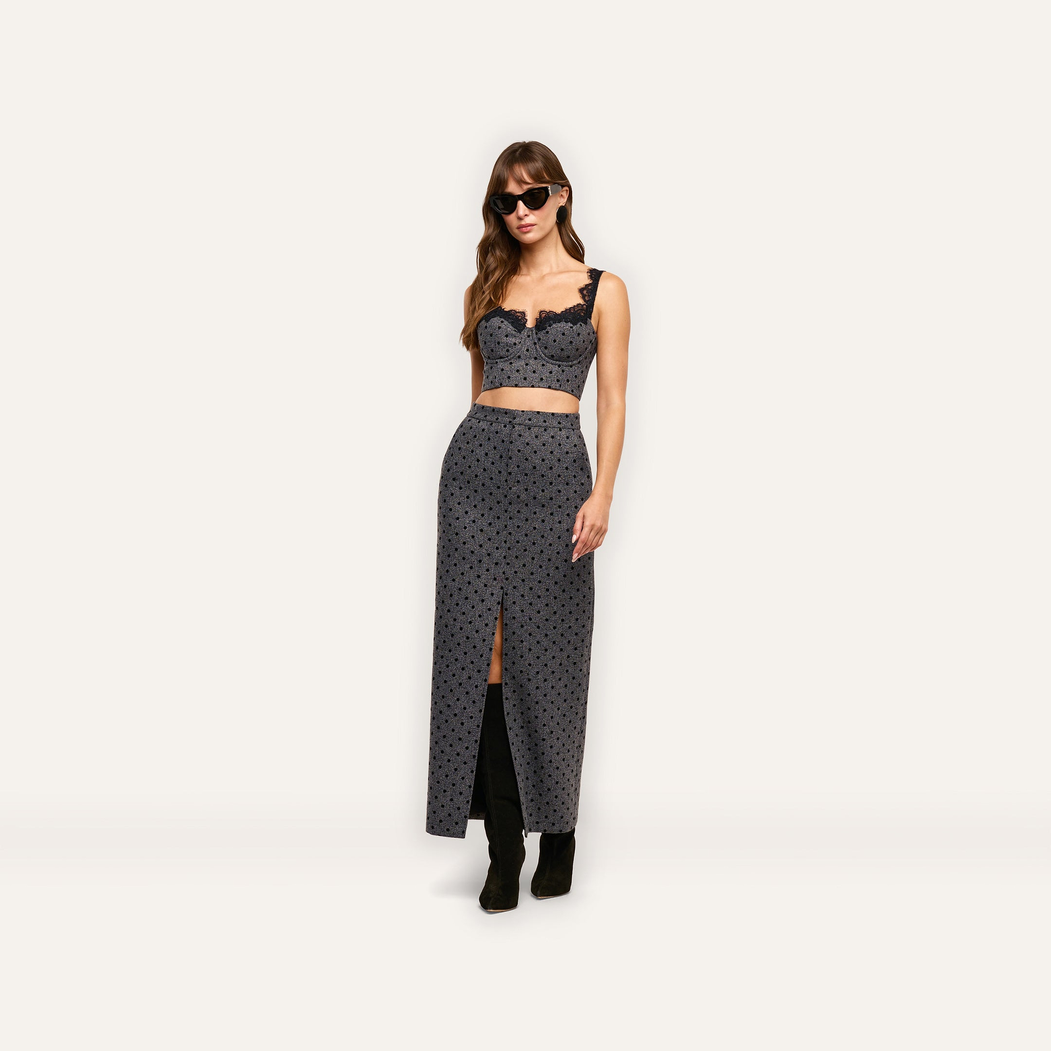 A person wearing an Oriane Bustier Top in gray with built-in underwire and black lace trim, paired with a matching high-waisted maxi skirt with a front slit, black sunglasses, and knee-high boots.