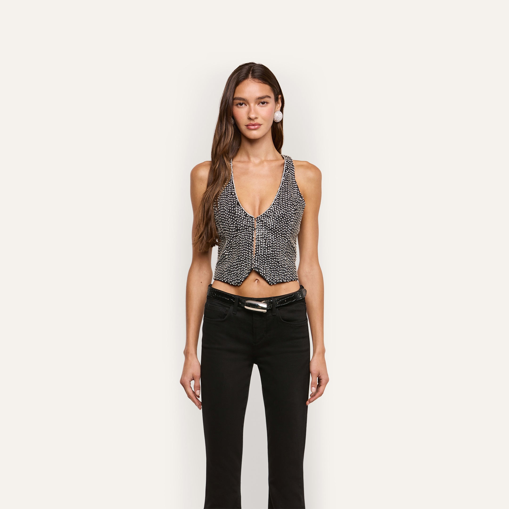 A person wearing a Nixe Embellished Vest, a cropped piece featuring a deep V-neck, front button closure, and a hand-embellished surface of reflective sequins and crystals, paired with black jeans.