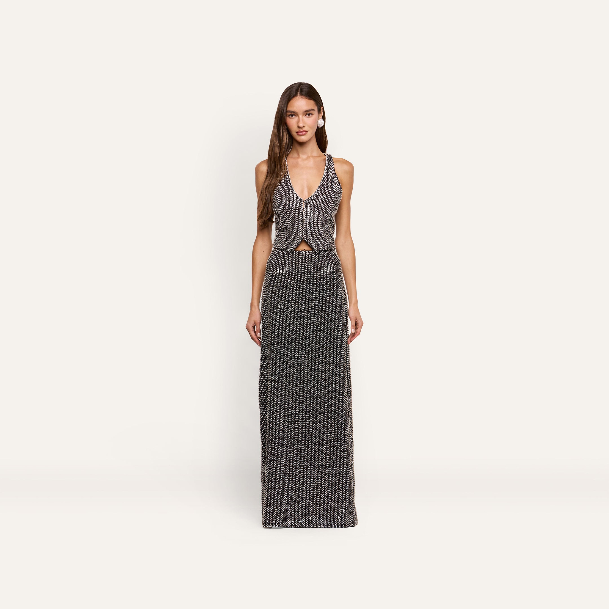 A woman wears a Nixe Embellished Vest, a cropped, hand-embellished piece with a deep V-neck, paired with a matching floor-length skirt, both covered in silver sequins and crystals.