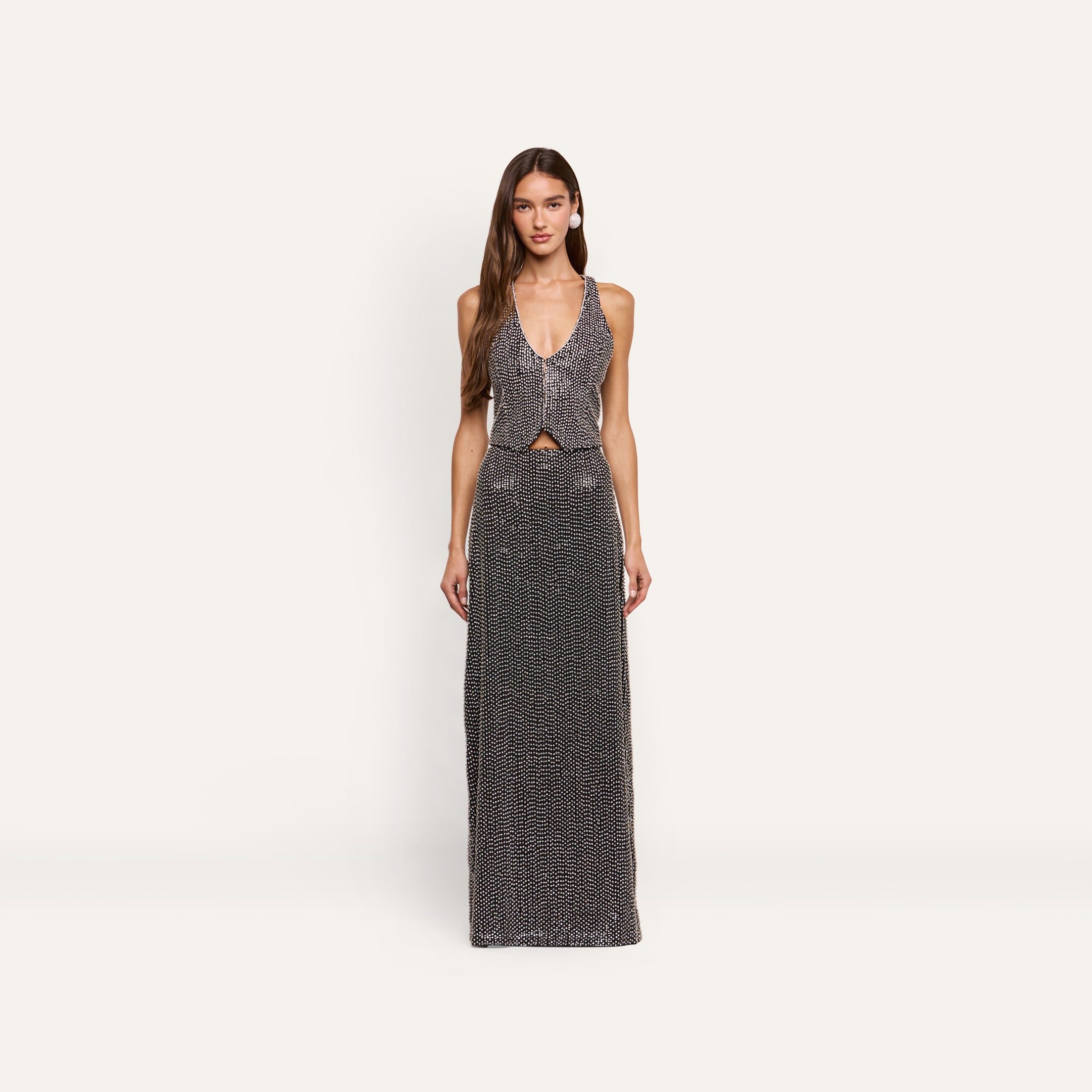 A woman wears a Nixe Embellished Vest, a cropped, hand-embellished piece with a deep V-neck, paired with a matching floor-length skirt, both covered in silver sequins and crystals.