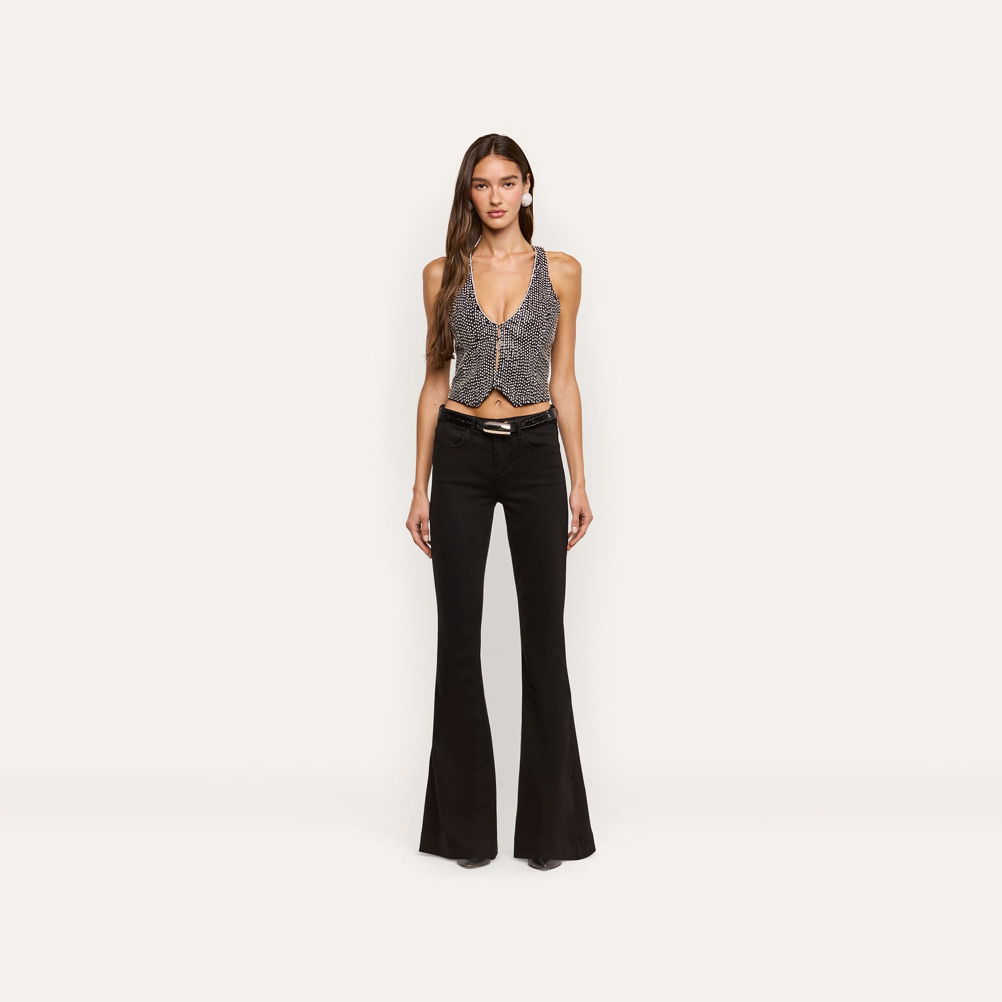 A person wears a hand-embellished Nixe Embellished Vest featuring a cropped, deep V-neck silhouette with small white dots, paired with black flare jeans and a black belt.