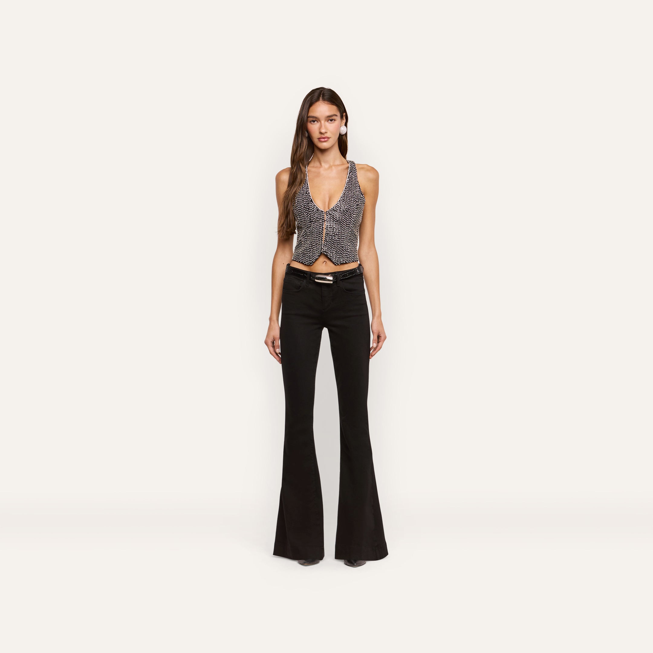 A person wears a hand-embellished Nixe Embellished Vest featuring a cropped, deep V-neck silhouette with small white dots, paired with black flare jeans and a black belt.