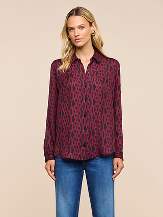 A woman wearing the Nina Silk Blouse—a maroon patterned, button-down silk top—paired with blue jeans stands against a beige background, looking directly at the camera with a neutral expression. The relaxed fit of the blouse easily transitions into a polished workplace ensemble.