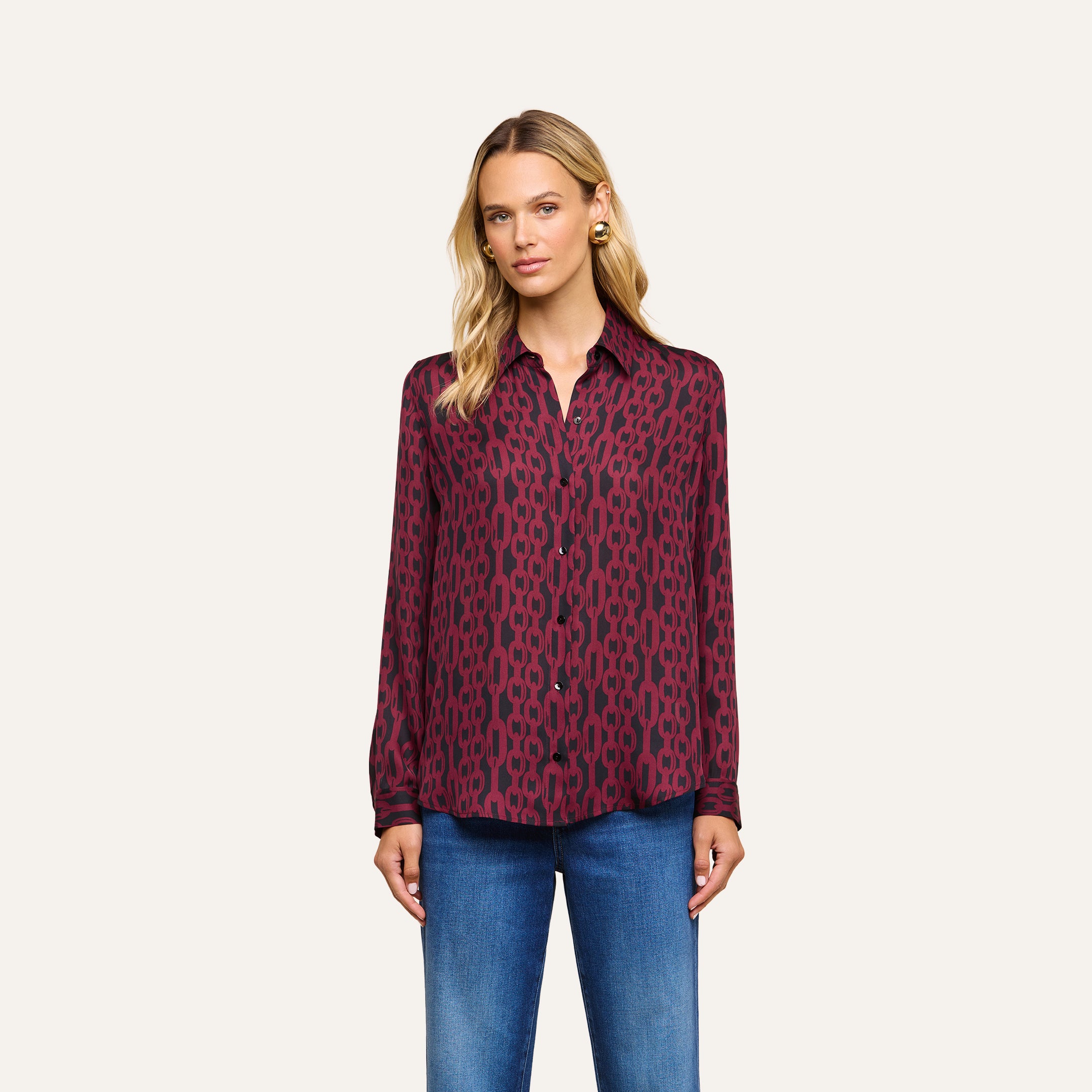 A woman stands against a plain, light background, wearing the Nina Silk Blouse—a relaxed fit, long-sleeve, patterned burgundy silk button-down shirt with dark designs—paired with blue jeans.