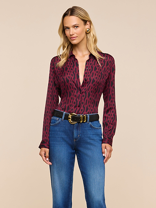 A woman stands against a beige background, looking forward with a slight smile while wearing the Nina Silk Blouse in a relaxed fit burgundy pattern, paired with blue jeans and a black belt featuring a gold buckle.