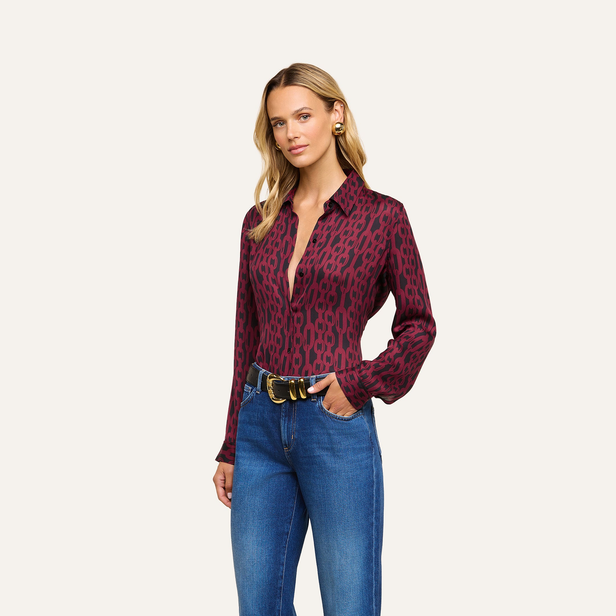 A woman wearing the Nina Silk Blouse—a maroon patterned, button-down top—paired with blue jeans and a black belt, stands against a plain light background, looking at the camera with a slight smile.