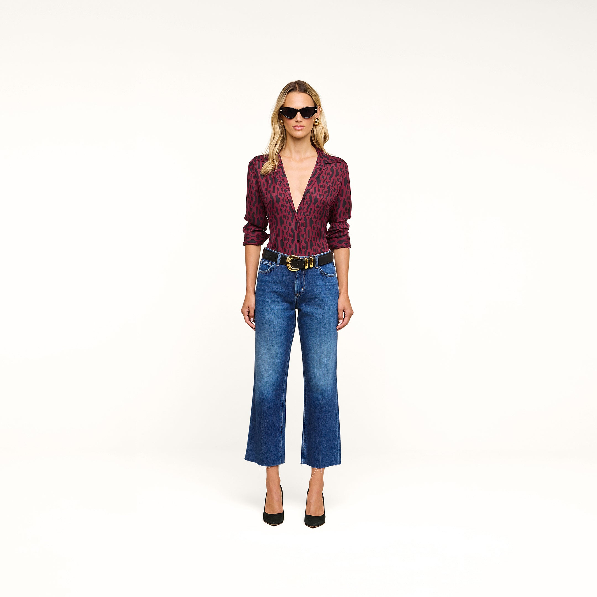 Woman in sunglasses wearing the Nina Silk Blouse—a relaxed fit, red patterned top—styled with blue cropped jeans, a black belt, and black high heels stands against a plain white background.
