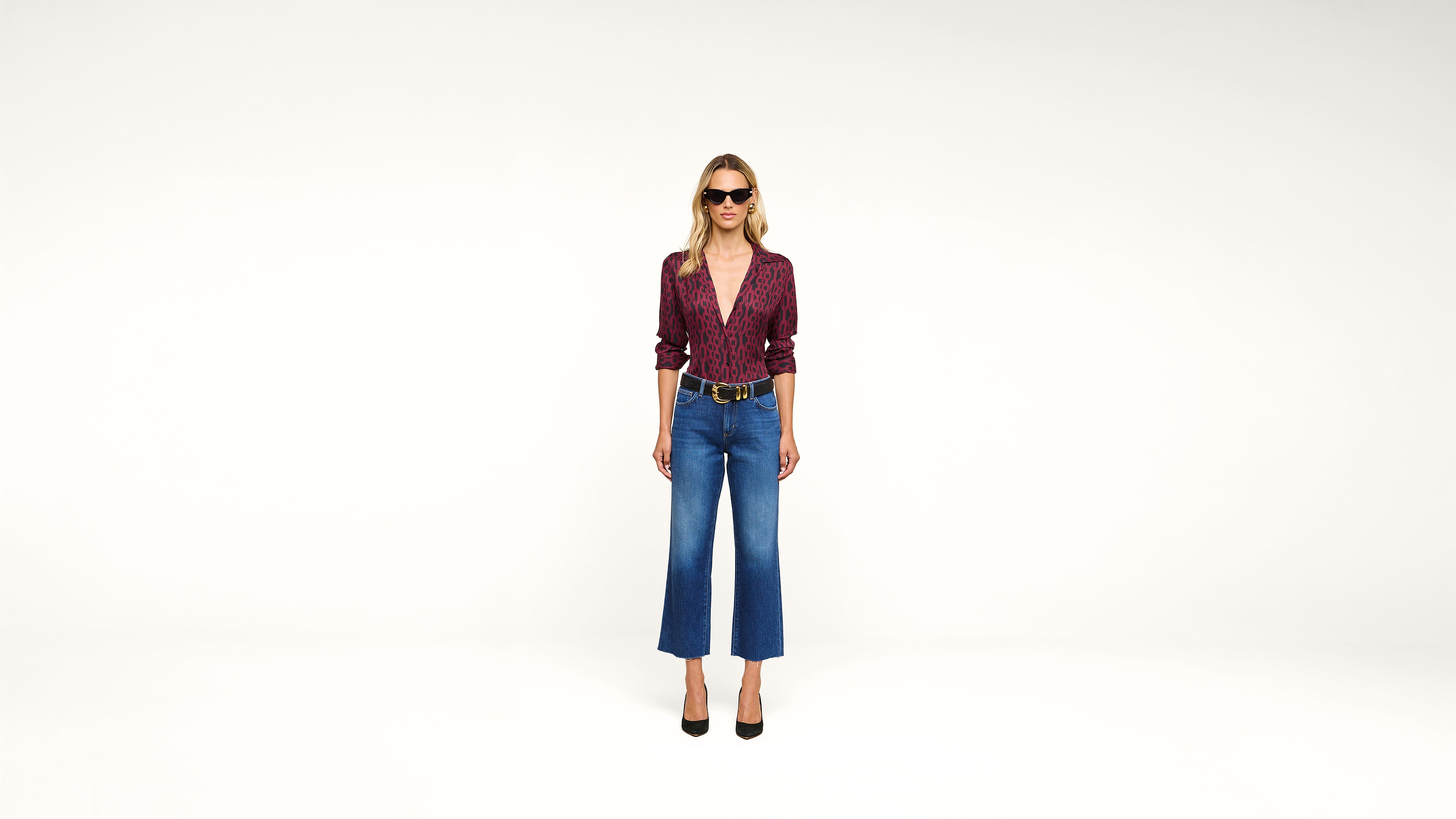 A woman wearing sunglasses, a patterned maroon blouse, blue high-waisted cropped jeans, and black heels stands against a plain white background.