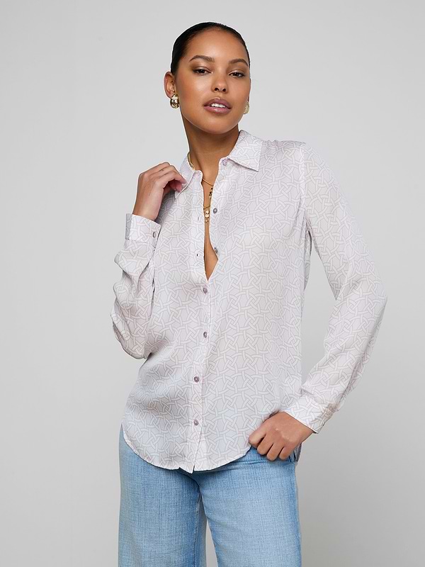 A woman poses confidently in the Nina Silk Blouse, a relaxed-fit geometric print button-up with the top buttons undone, paired with light blue jeans. She wears gold earrings and a necklace. Her dark hair is pulled back against a plain light gray background.