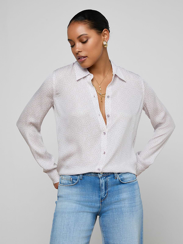 A woman with her hair pulled back wears the relaxed fit Nina Silk Blouse with a geometric print and light blue jeans. She stands with hands in pockets, looking down slightly, accessorized with gold earrings and layered necklaces.