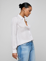 A woman stands in profile against a plain background, wearing the relaxed fit Nina Silk Blouse—a light-colored, long-sleeved button-up shirt—paired with light blue jeans. Her hair is styled in a neat bun and she gazes downward.