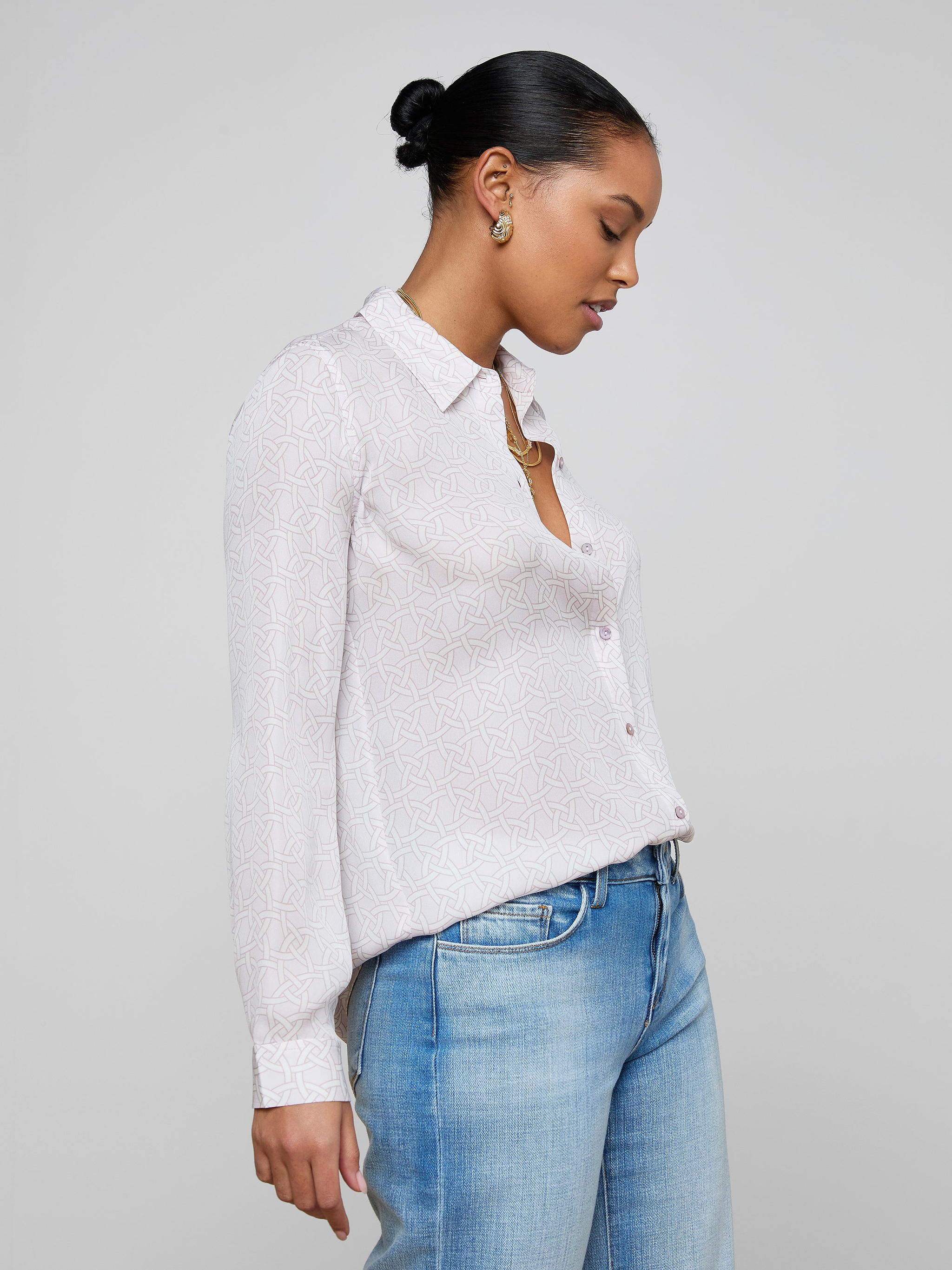 A woman stands in profile against a plain background, wearing the relaxed fit Nina Silk Blouse—a light-colored, long-sleeved button-up shirt—paired with light blue jeans. Her hair is styled in a neat bun and she gazes downward.