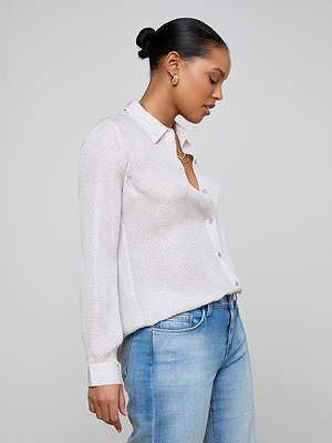 A woman with dark hair in a low bun wears the Nina Silk Blouse—white with a geometric print—in a relaxed fit and light blue jeans. She accessorizes with gold hoop earrings and a necklace, looking down neutrally against a plain light background.