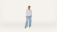 A woman stands against a plain white background, wearing a white button-up shirt, light blue wide-leg jeans, and tan sandals, looking off to the side.