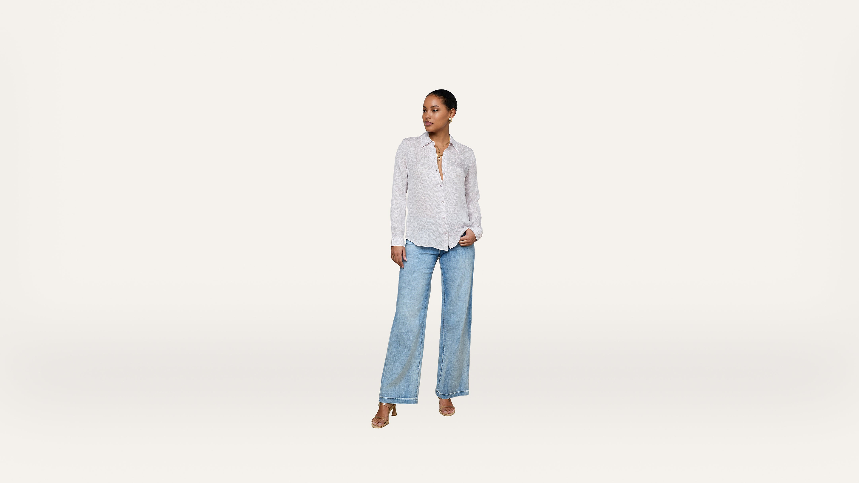 A woman stands against a plain white background, wearing a white button-up shirt, light blue wide-leg jeans, and tan sandals, looking off to the side.