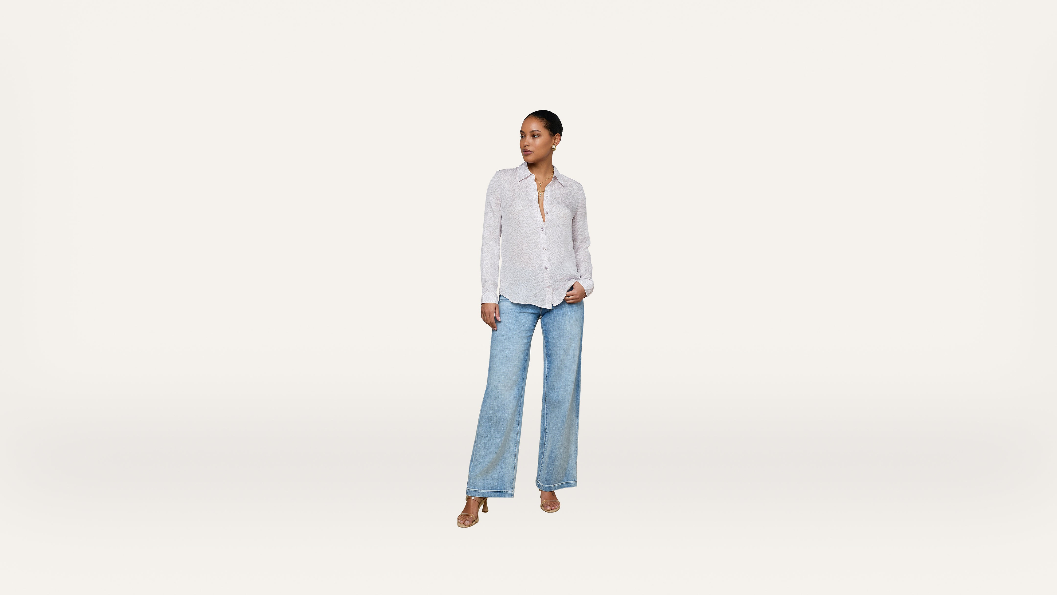 A woman stands against a plain white background, wearing a white button-up shirt, light blue wide-leg jeans, and tan sandals, looking off to the side.