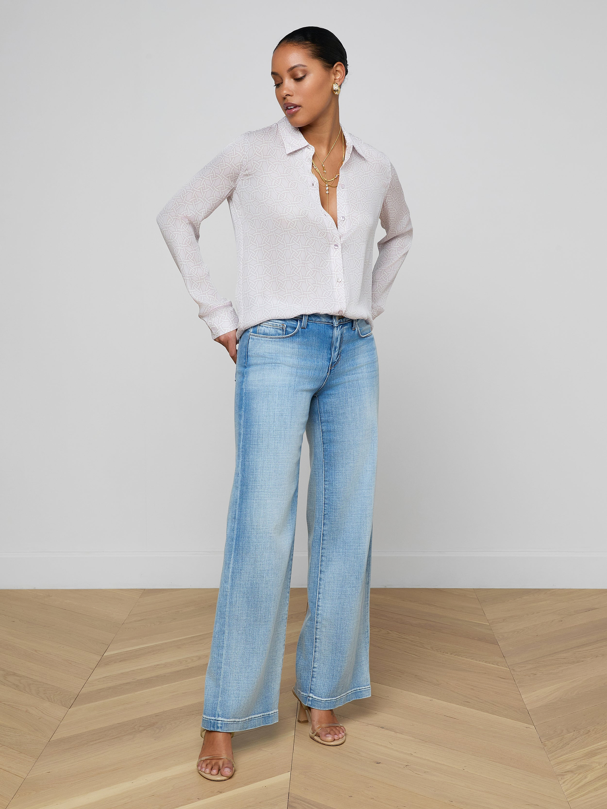 A woman stands against a plain white background, wearing the Nina Silk Blouse, light blue wide-leg jeans, and heeled sandals. She looks slightly to her left with one hand on her hip.
