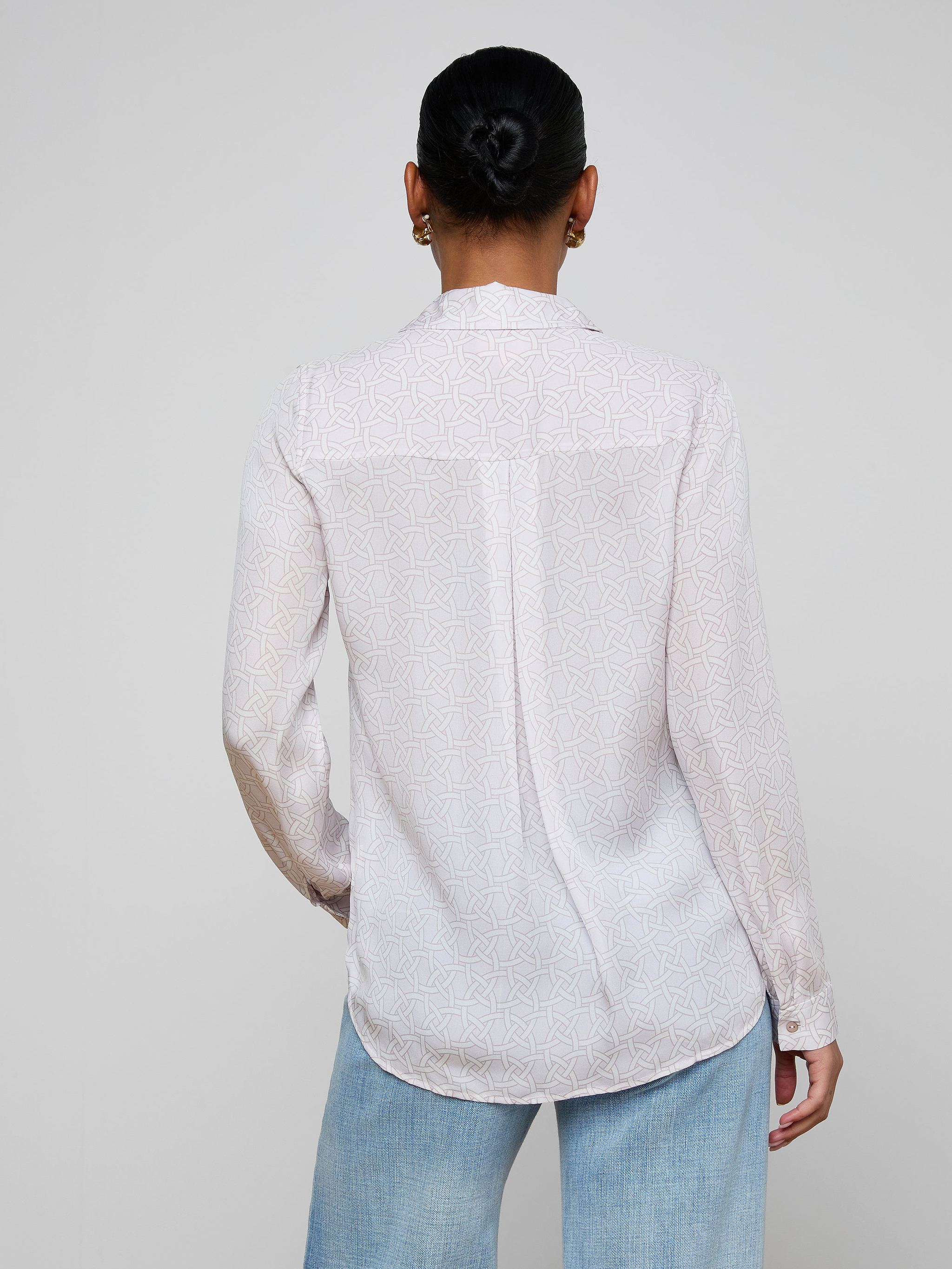 A person stands facing away, wearing the Nina Silk Blouse—a white silk button down with a subtle geometric print—paired with relaxed fit light blue jeans. Their hair is styled in a neat bun.