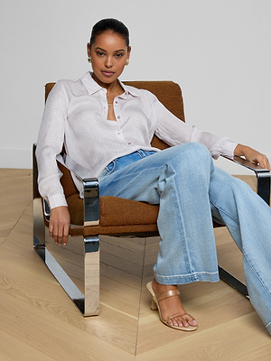A woman sits confidently in a modern brown and chrome chair, wearing the Nina Silk Blouse with blue wide-leg jeans and beige heeled sandals, in a minimal room with wood flooring.