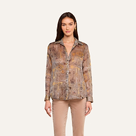 Una mujer aparece de pie mirando hacia delante, con la blusa de seda «Nina» —una blusa de corte holgado, de manga larga y con botones, en tonos marrones y beige— combinada con unos pantalones marrón claro, sobre un fondo liso de color beige.