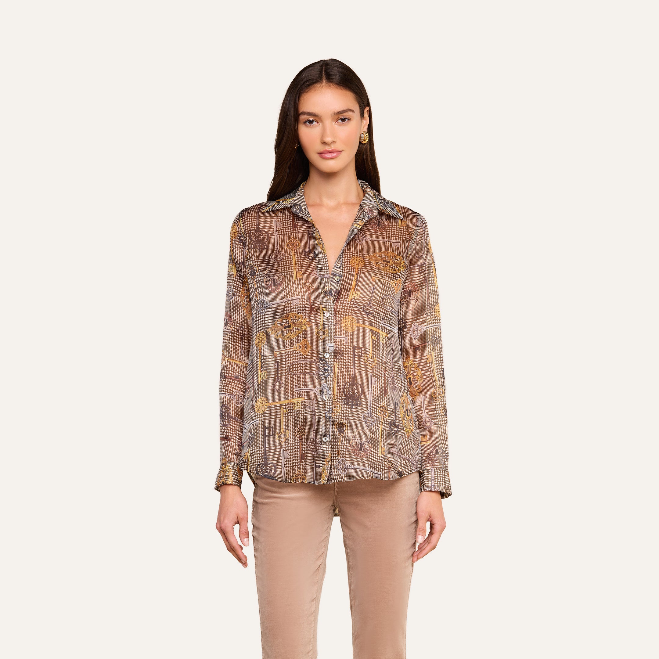 Una mujer aparece de pie mirando hacia delante, con la blusa de seda «Nina» —una blusa de corte holgado, de manga larga y con botones, en tonos marrones y beige— combinada con unos pantalones marrón claro, sobre un fondo liso de color beige.