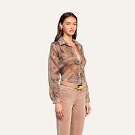 Una mujer posa frente a un fondo beige, vestida con la blusa Nina Silk —una camisa abotonada de corte holgado con estampado de pata de gallo marrón— combinada con unos pantalones beige, un cinturón beige con hebilla dorada y unos pendientes dorados. Tiene el pelo largo y castaño y una expresión neutra.