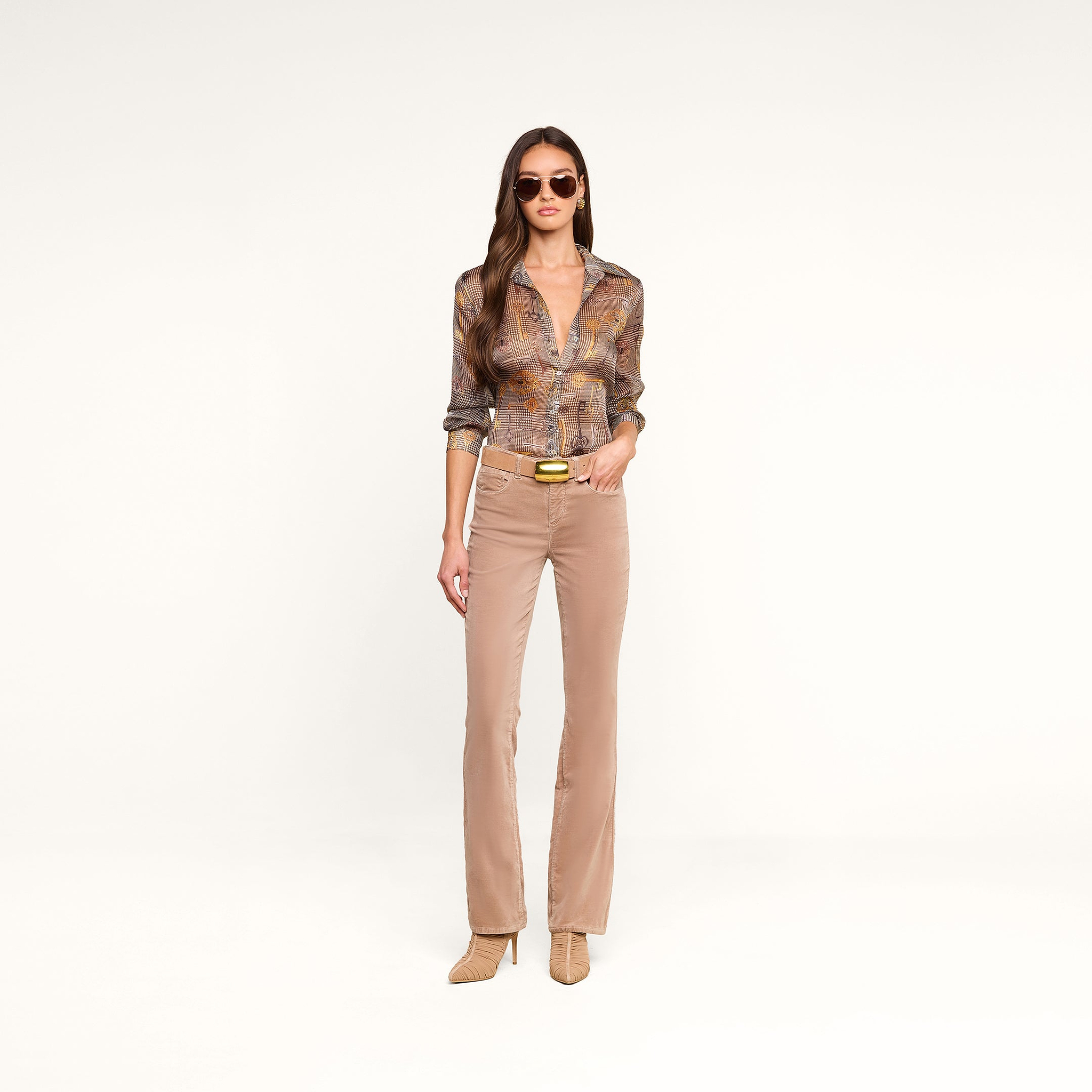 Una mujer posa frente a un fondo beige con gafas de sol, la blusa «Nina Silk» con estampado de pata de gallo marrón y corte holgado, pantalones acampanados beige con cinturón y botas de tacón. Lleva el pelo largo y castaño suelto y mira al frente con expresión neutra.