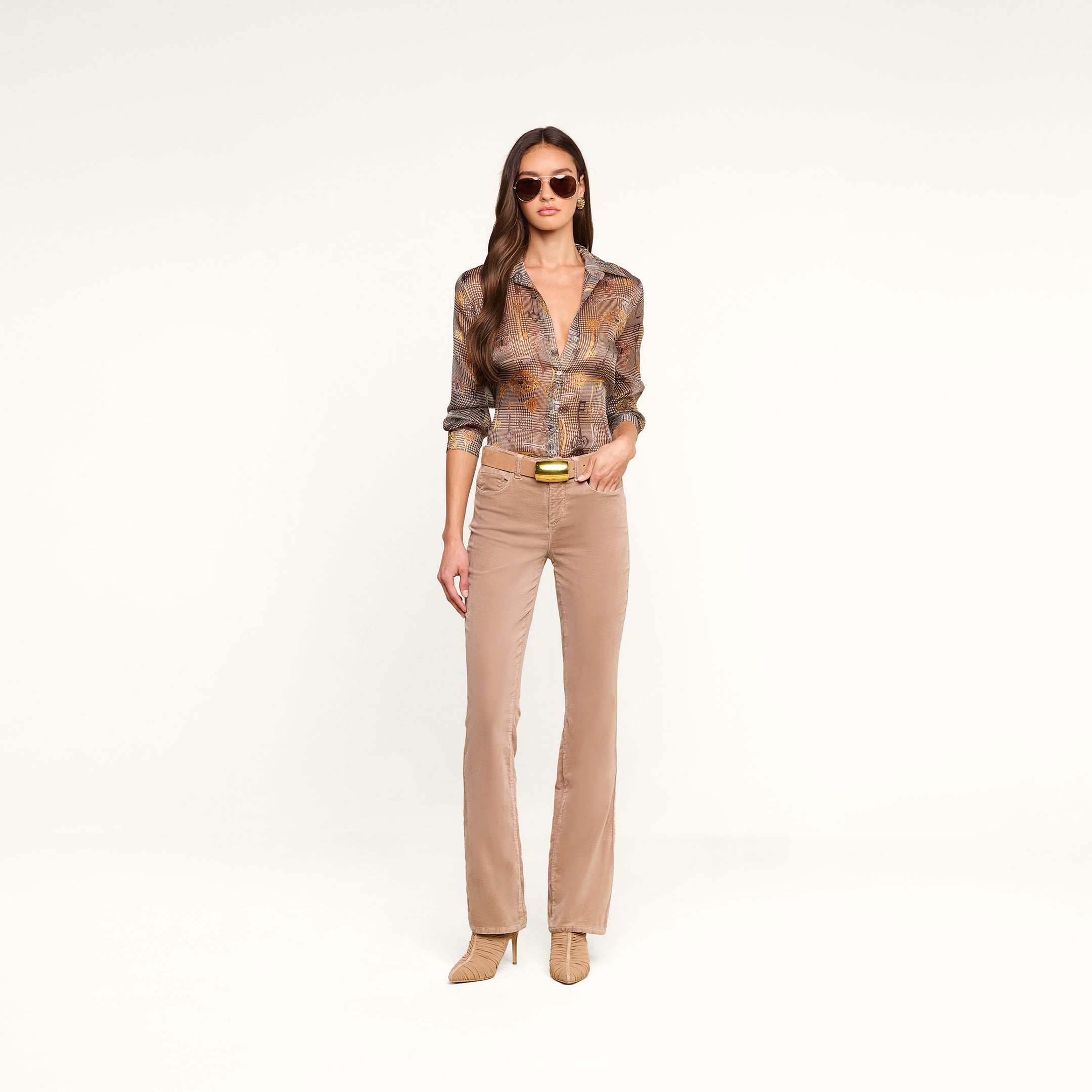 Una mujer posa frente a un fondo beige con gafas de sol, la blusa «Nina Silk» con estampado de pata de gallo marrón y corte holgado, pantalones acampanados beige con cinturón y botas de tacón. Lleva el pelo largo y castaño suelto y mira al frente con expresión neutra.