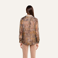 Se ve a una persona con el pelo largo y castaño, de espaldas, que lleva la blusa de seda «Nina» —una blusa de seda transparente con estampado y de corte holgado— combinada con unos pantalones beige, de pie frente a un fondo liso de color beige.