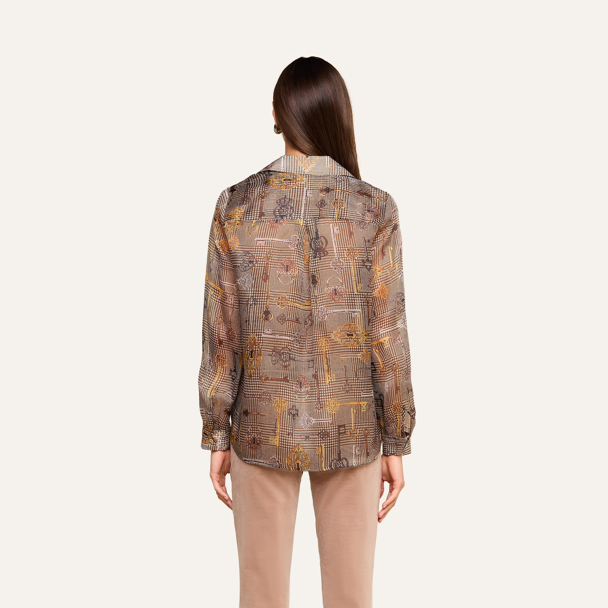 Se ve a una persona con el pelo largo y castaño, de espaldas, que lleva la blusa de seda «Nina» —una blusa de seda transparente con estampado y de corte holgado— combinada con unos pantalones beige, de pie frente a un fondo liso de color beige.