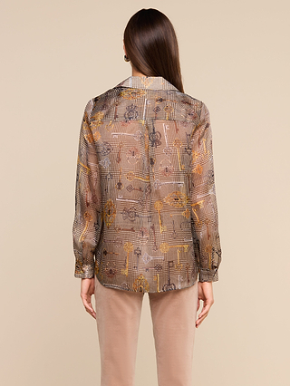 A person with long brown hair is shown from behind wearing the Nina Silk Blouse—a sheer, patterned silk shirt in a relaxed fit—paired with beige pants, standing against a plain beige background.