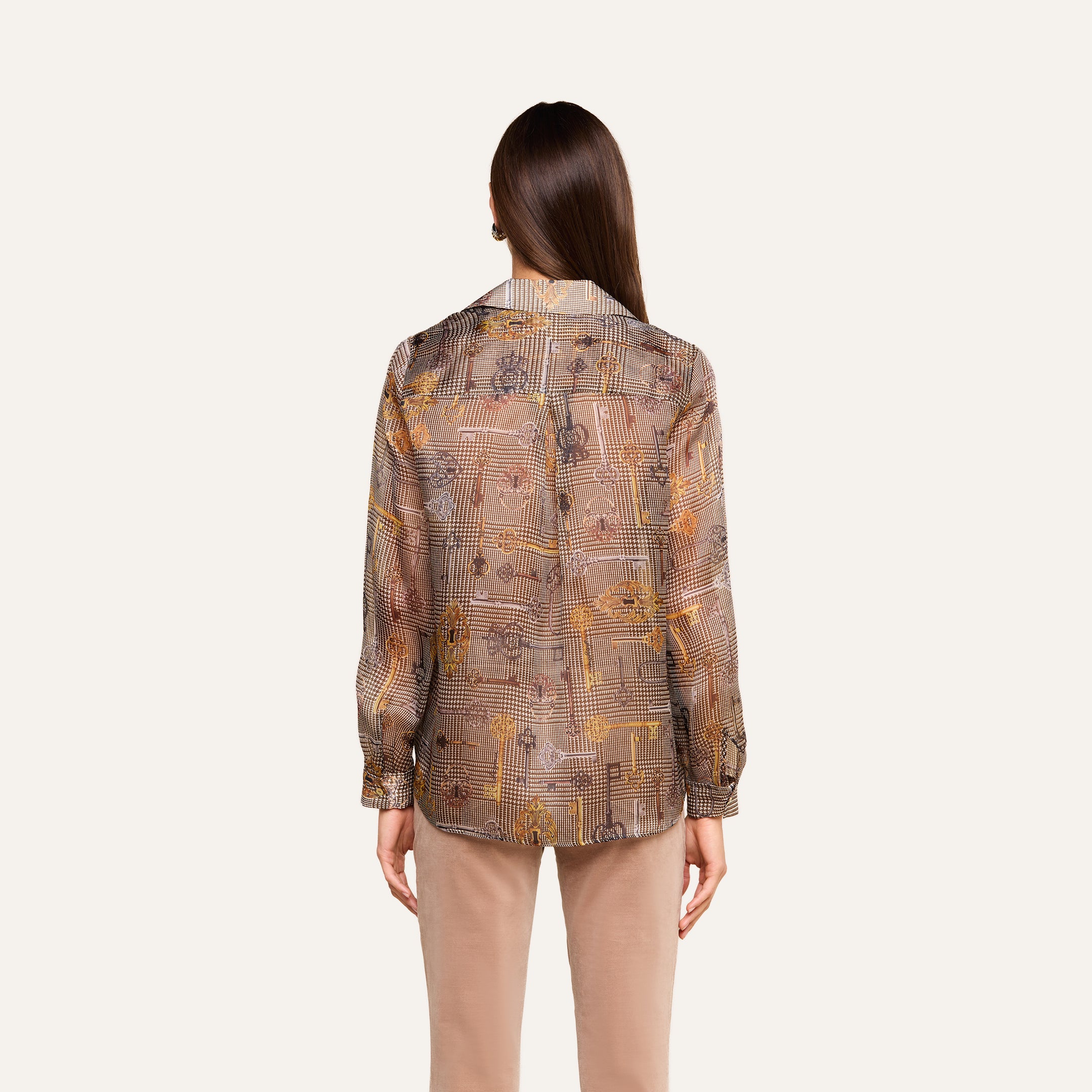 Se ve a una persona con el pelo largo y castaño, de espaldas, que lleva la blusa de seda «Nina» —una blusa de seda transparente con estampado y de corte holgado— combinada con unos pantalones beige, de pie frente a un fondo liso de color beige.