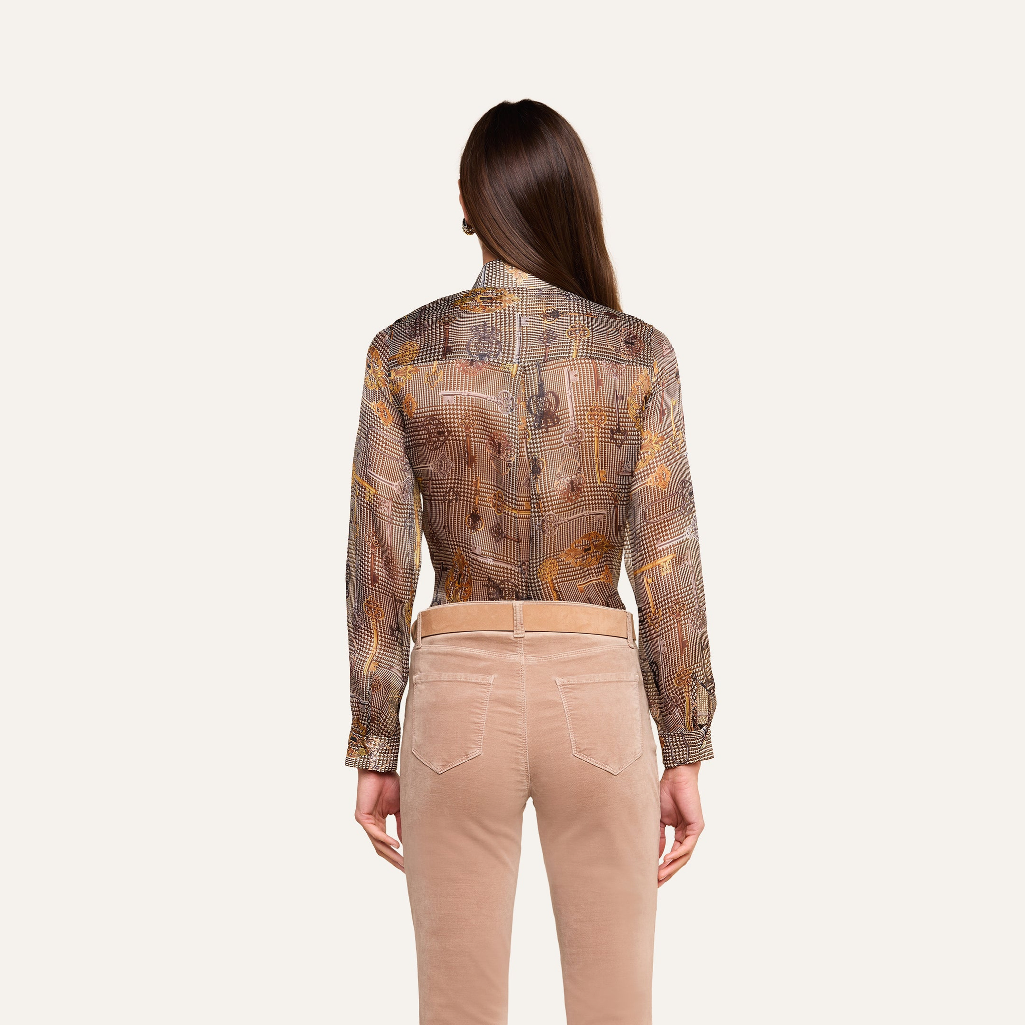 Se muestra a una persona con el pelo largo y castaño, vista de espaldas sobre un fondo neutro, que lleva la blusa Nina Silk —una blusa transparente con estampado y corte holgado— combinada con unos pantalones beige.