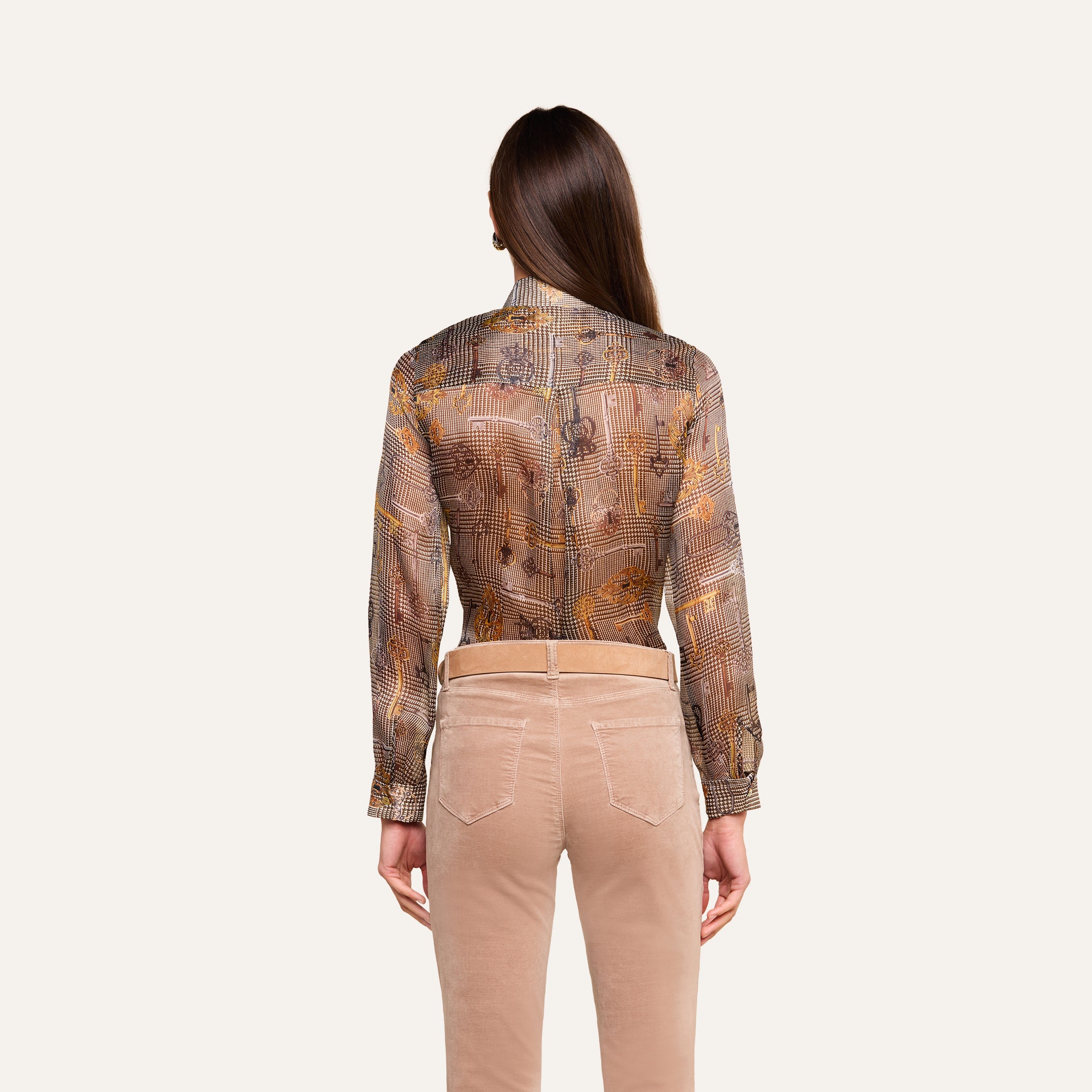 Se muestra a una persona con el pelo largo y castaño, vista de espaldas sobre un fondo neutro, que lleva la blusa Nina Silk —una blusa transparente con estampado y corte holgado— combinada con unos pantalones beige.