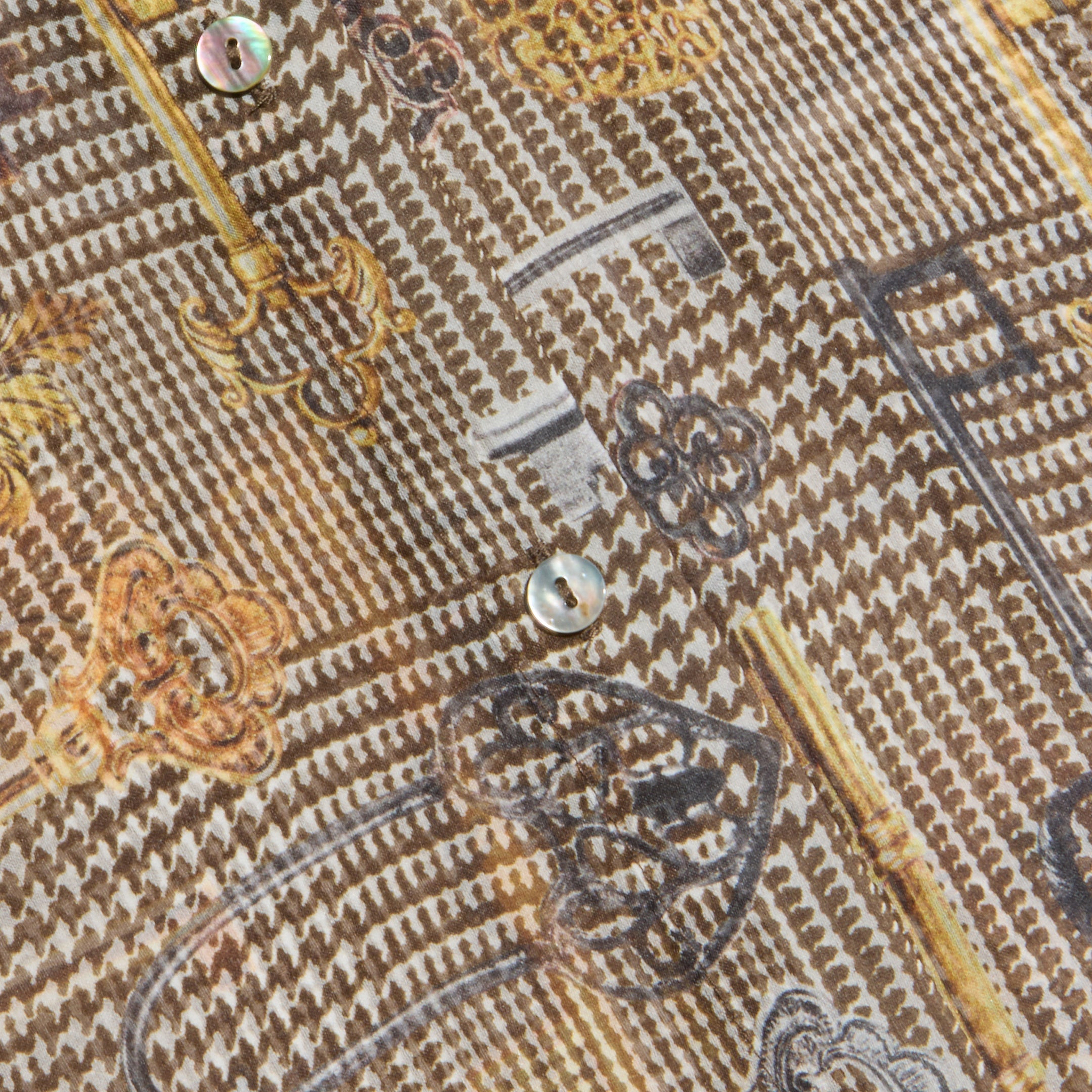 Close-up of the Nina Silk Blouse, showcasing a patterned design with vintage keys in gold, black, and gray tones. The fabric is adorned with small iridescent buttons and features a textured, woven appearance.