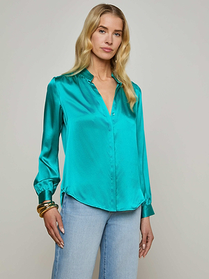 A woman with long blonde hair wears the Bianca Blouse—a shiny teal silk top with a relaxed fit and long sleeves—paired with light blue jeans and gold bangles, standing against a plain light background.