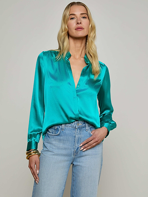 A woman with long blonde hair models the Bianca Blouse—a shiny teal silk top with a relaxed fit—paired with light blue jeans, standing with one hand in her pocket against a plain light background.