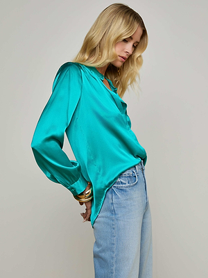 A person with long blonde hair wears the Bianca Blouse in bright teal silk and light blue high-waisted relaxed jeans, standing sideways against a plain background.