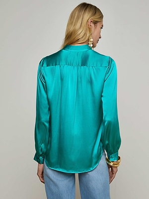 A woman with long blonde hair, wearing the Bianca Blouse—a teal silk top with a relaxed fit—and light blue jeans, stands facing away from the camera. She accessorizes with large gold earrings and bangles.