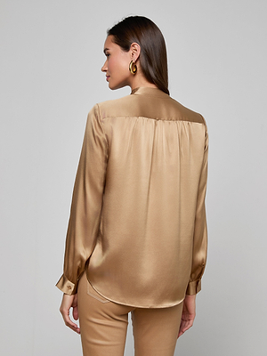 A woman with straight brown hair faces away, wearing the Bianca Silk Blouse in gold. The long-sleeve charmeuse blouse has a relaxed silhouette and subtle sheen, paired with matching pants and large hoop earrings.