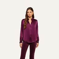 A woman stands facing forward, wearing the Bianca Silk Blouse—a shiny, long-sleeved burgundy silk charmeuse top—paired with matching pants. She has long brown hair and a neutral expression, posed against a plain off-white background.