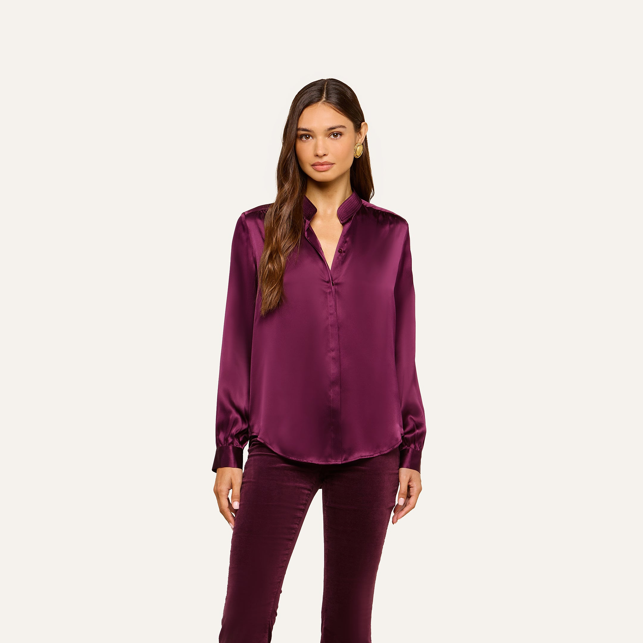 A woman stands facing forward, wearing the Bianca Silk Blouse—a shiny, long-sleeved burgundy silk charmeuse top—paired with matching pants. She has long brown hair and a neutral expression, posed against a plain off-white background.