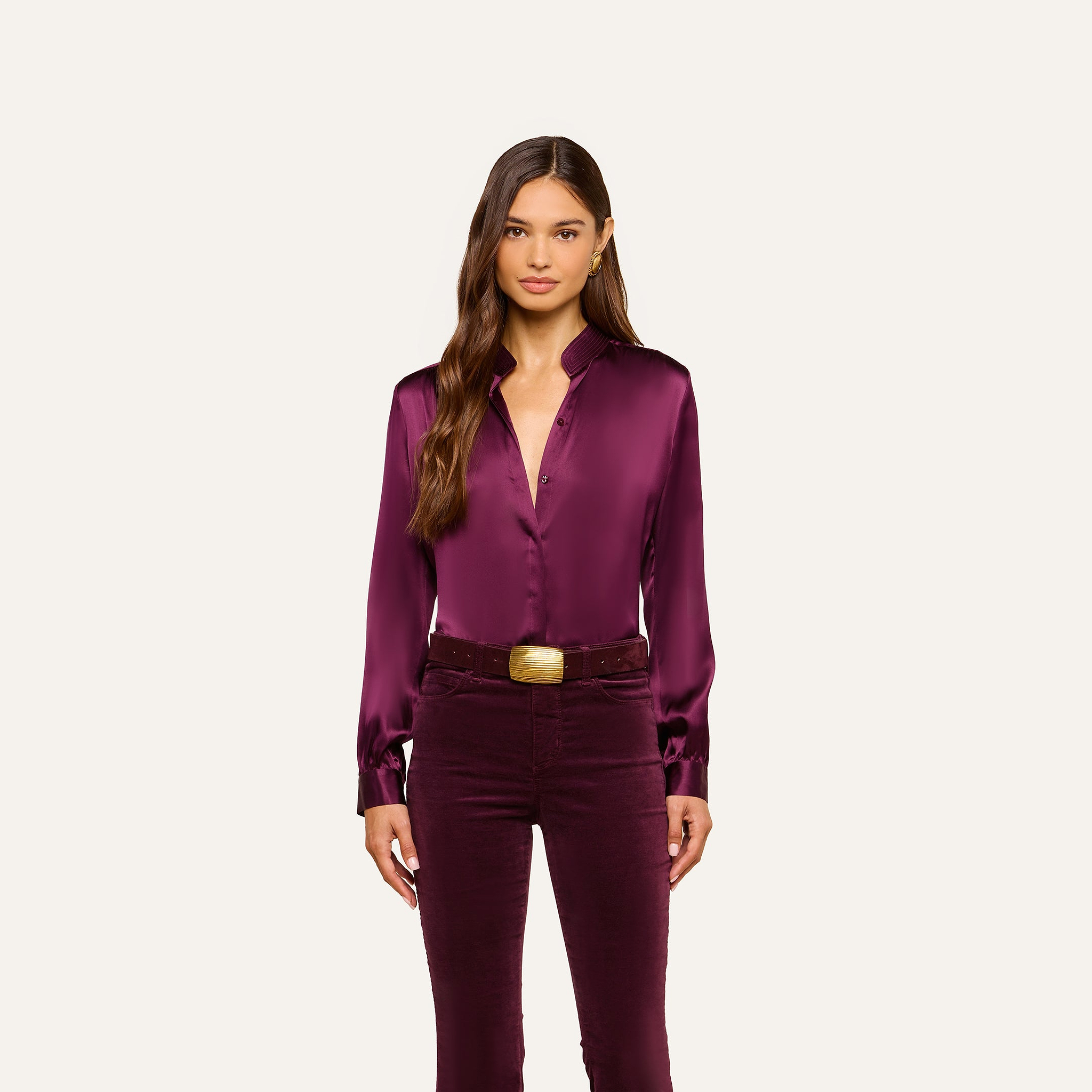 Wearing the Bianca Silk Blouse in deep purple, a woman stands facing forward with her arms relaxed at her sides. She pairs the blouse with matching pants and accessorizes with a wide brown belt featuring a large gold buckle. Her long brown hair frames her neutral expression.